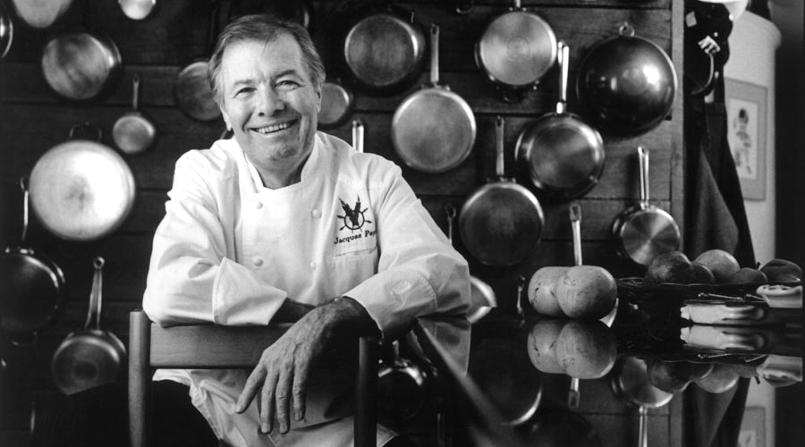 Black and white photo of Jacques Pepin standing in front of a wall with pans hanging on it