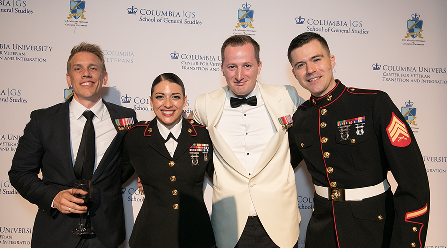 Four guests pose in front of a step-and-repeat, all displaying mini-medals for their service, and 2 in military dress