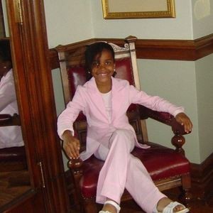 Nine-year-old Justice Betty sitting in a chair in a pink pant suit.