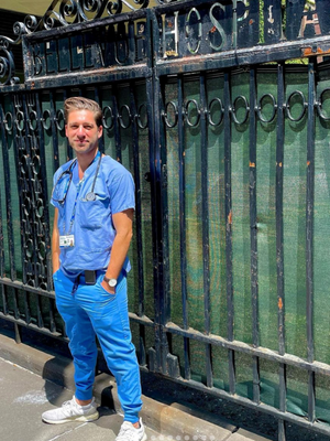 Michael Natter ‘11PBPM in scrubs standing in front of the Bellevue Hospital gate