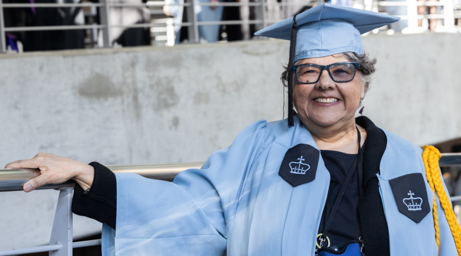 Columbia GS graduate Lisa Delgado Smith '25GS poses beside a railing in Lerner Hall wearing a light blue Columbia graduation gown and cap.