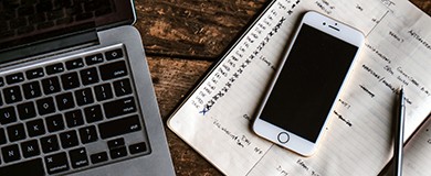 laptop next to a notebook and pen with an iPhone on top