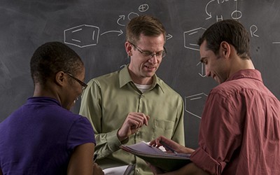 A professor and two students go over notes in front of a chalkboard