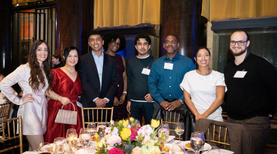 Eight people dressed in formal wear lined up behind a table in Low Library, smiling and looking into the camera. 