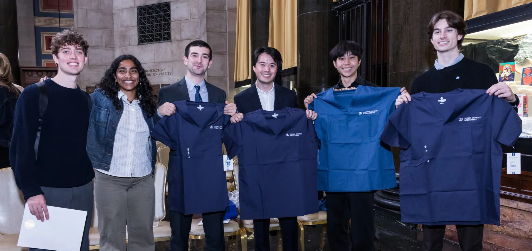 Postbac Premed Program graduates holding up Postbac Premed-branded scrubs in Low Library