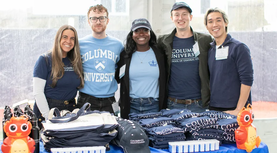A group of GS staff and alumni pose in front of a table of Columbia GS swag at homecoming weekend.