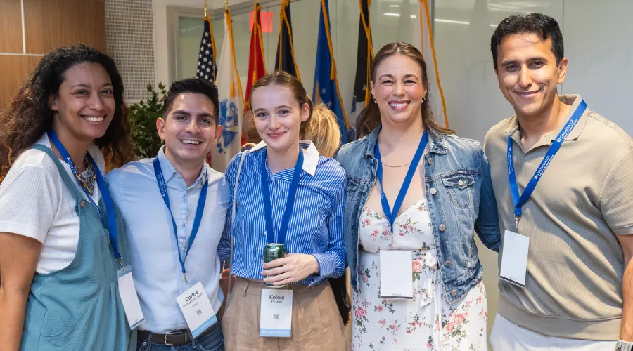 A group of five smiling Columbia GS alumni wearing name lanyards pose for a photo during Columbia Reunion Weekend. Flags are displayed in the background.