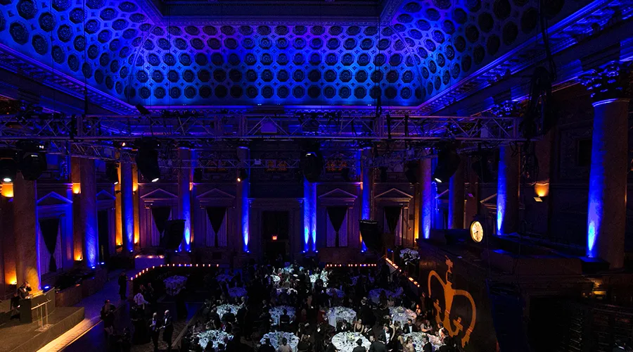 Picture of a large, blue-lit room with high, ornate ceilings looking down at round tables with people sitting