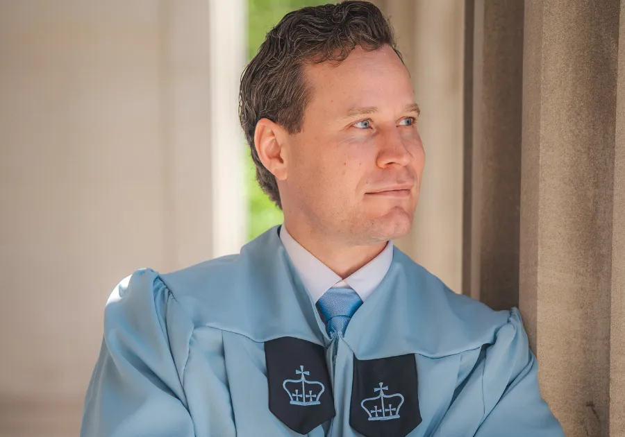Brian Williams '25GS in a Columbia blue graduation gown looks off into the distance.