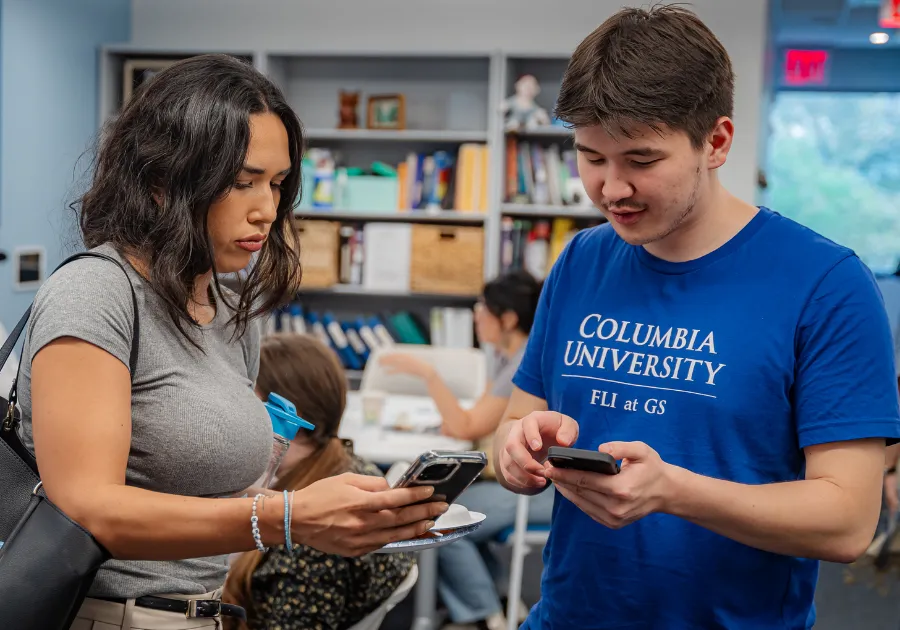 Two students sharing information from their mobile phones in a study area with a bookshelf in the background.