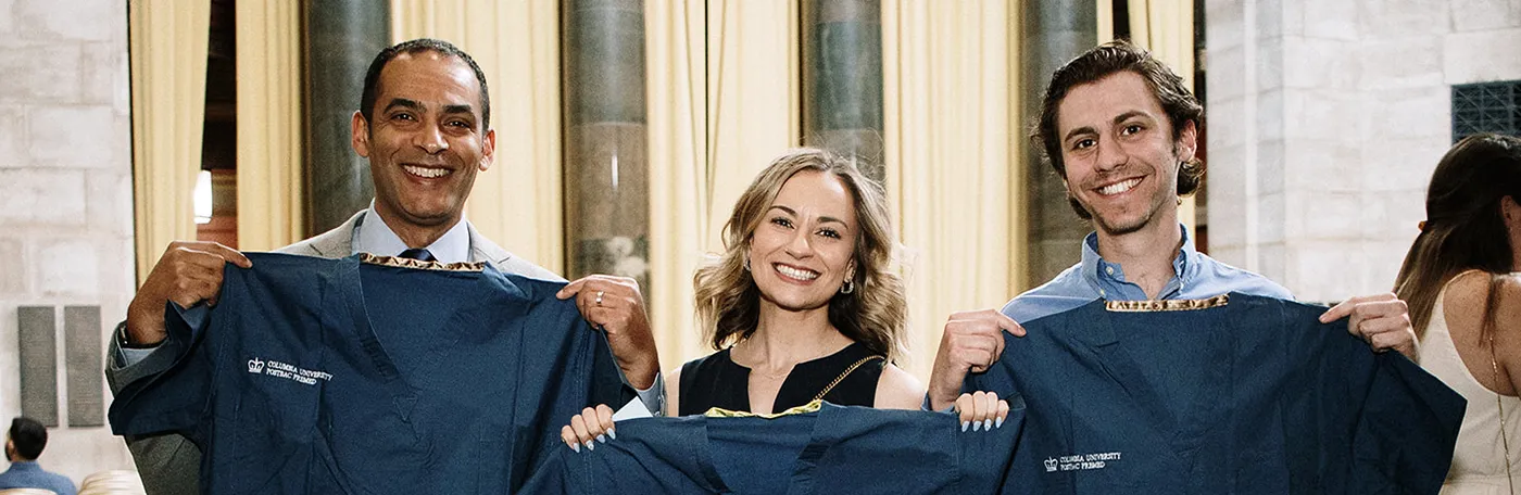 Three Postbac Premed Program graduates holding up Postbac Premed-branded scrubs in Low Library